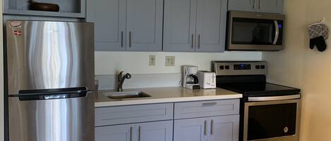 Una cocina compacta con gabinetes grises, un refrigerador de acero inoxidable, un microondas, una cafetera y un horno.