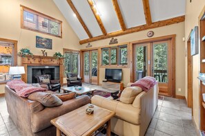 A cozy living room with leather sofas, a fireplace, a television, and large windows offering a view of the outdoors.