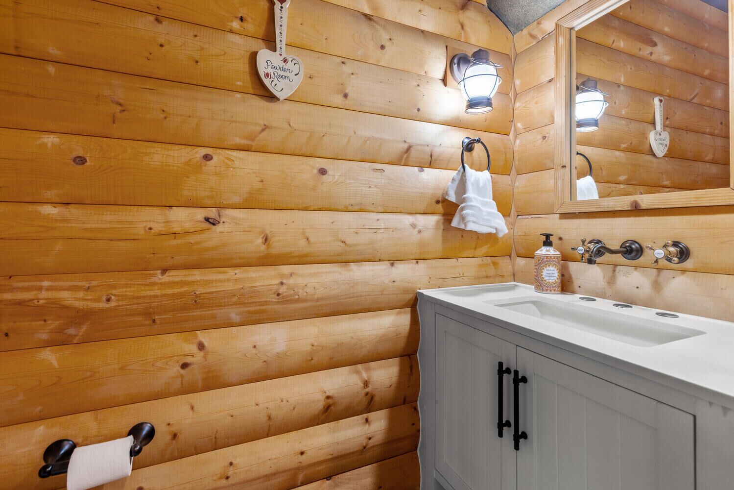 Un bagno in stile baita con pareti in legno, un lavandino bianco, uno specchio e un portascopevoli.