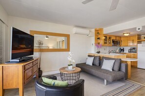A modern living room with a sofa, coffee table, and television. The kitchen is visible in the background with wooden cabinets and a microwave.