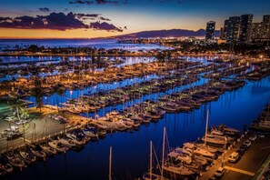 A marina with numerous boats docked, a city skyline in the background, and a sunset over the water.