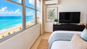 A bedroom with a large window overlooking a beach, a flat-screen TV, and a dark wooden dresser.