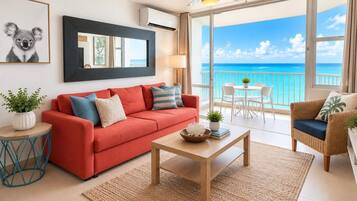 A room with a red sofa, a wooden coffee table, a wicker chair, and a view of the ocean through large windows.