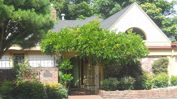 Une maison avec une façade en briques, une cheminée et un jardin devant, agrémenté de différentes plantes et d’un arbre.