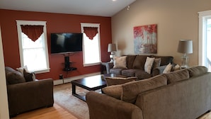 A living room with a large flat-screen TV, two windows with curtains, a sofa set, a coffee table, and a painting on the wall. - GORGEOUS AND MODERN describes this 6 bedroom home . (Warrensburg)