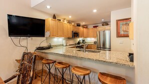 A modern kitchen with wooden cabinets, a granite countertop, a stainless steel refrigerator, and a flat-screen TV mounted on the wall. - Spacious 4BR Townhome | 2 Living Rooms and Walk to Ski Area! (Steamboat Springs)