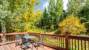 A wooden deck with a table and chairs, surrounded by trees and a view of a house in the background. - Spacious 4BR Townhome | 2 Living Rooms and Walk to Ski Area! (Steamboat Springs)