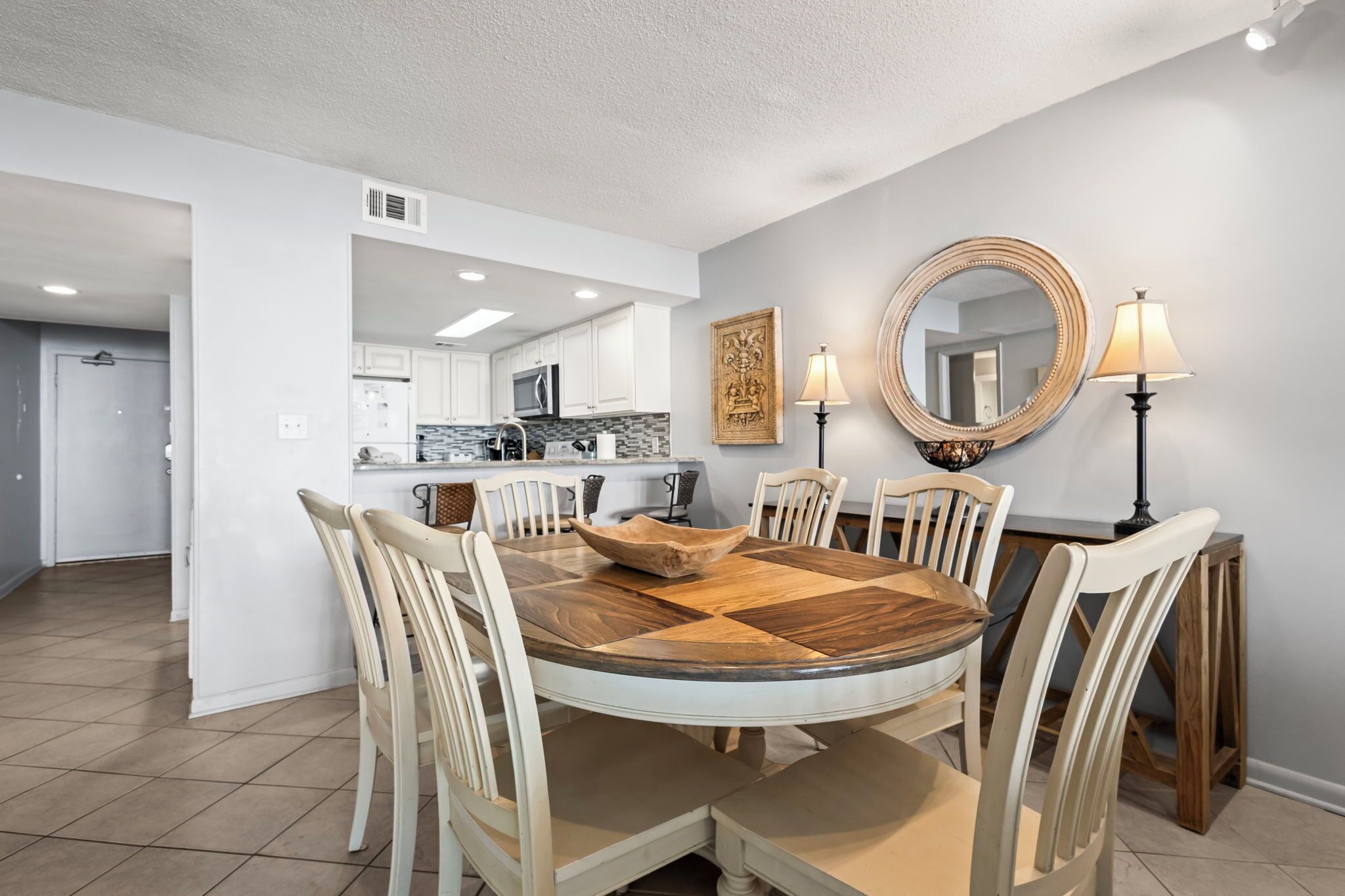 Un coin repas comprenant une table ronde en bois, des chaises blanches, un grand miroir et deux lampadaires.