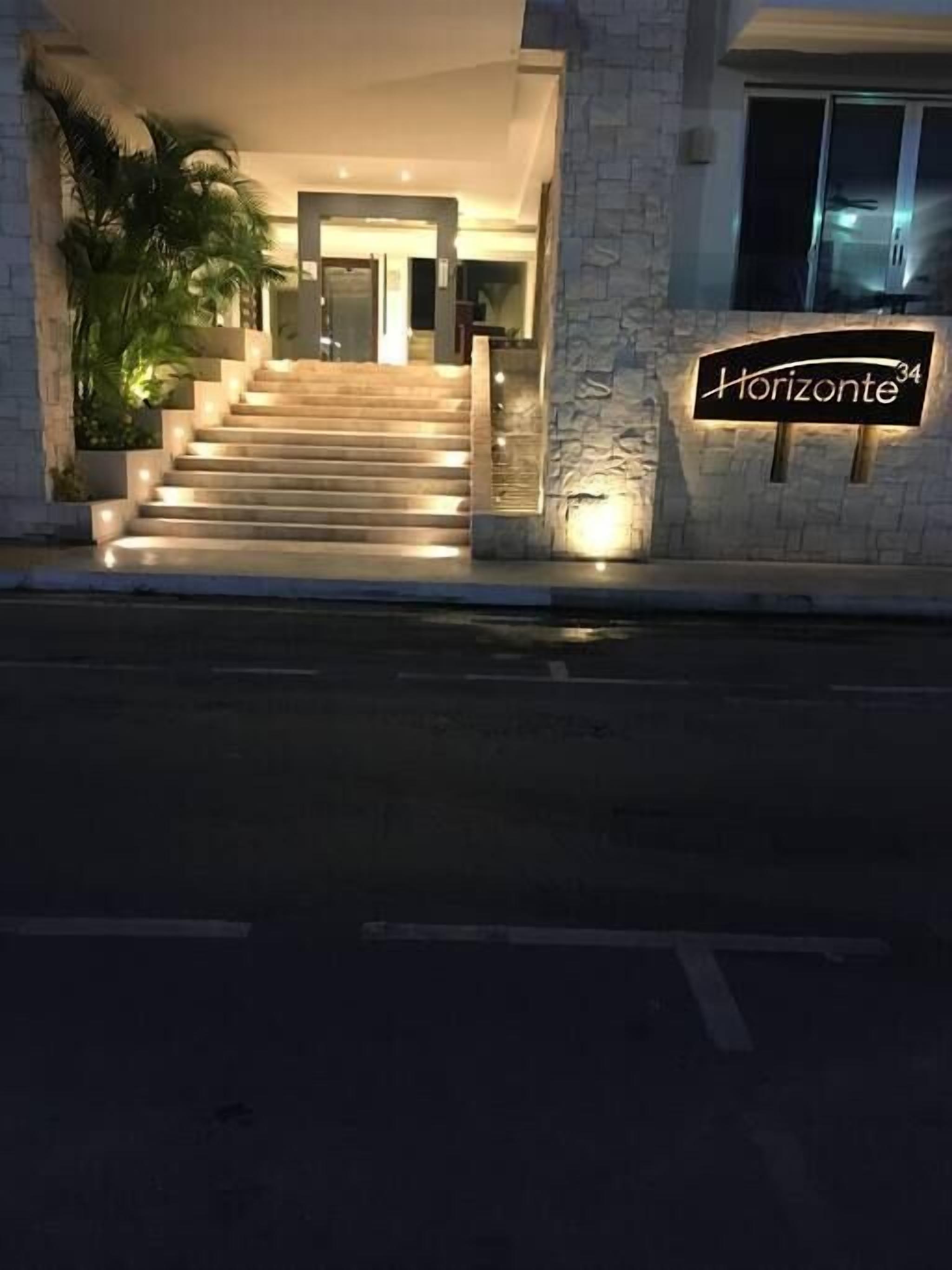 L’entrée d’un hôtel, avec un escalier menant au hall, une enseigne indiquant « Horizonte », et un pot de fleurs placé sur la gauche.