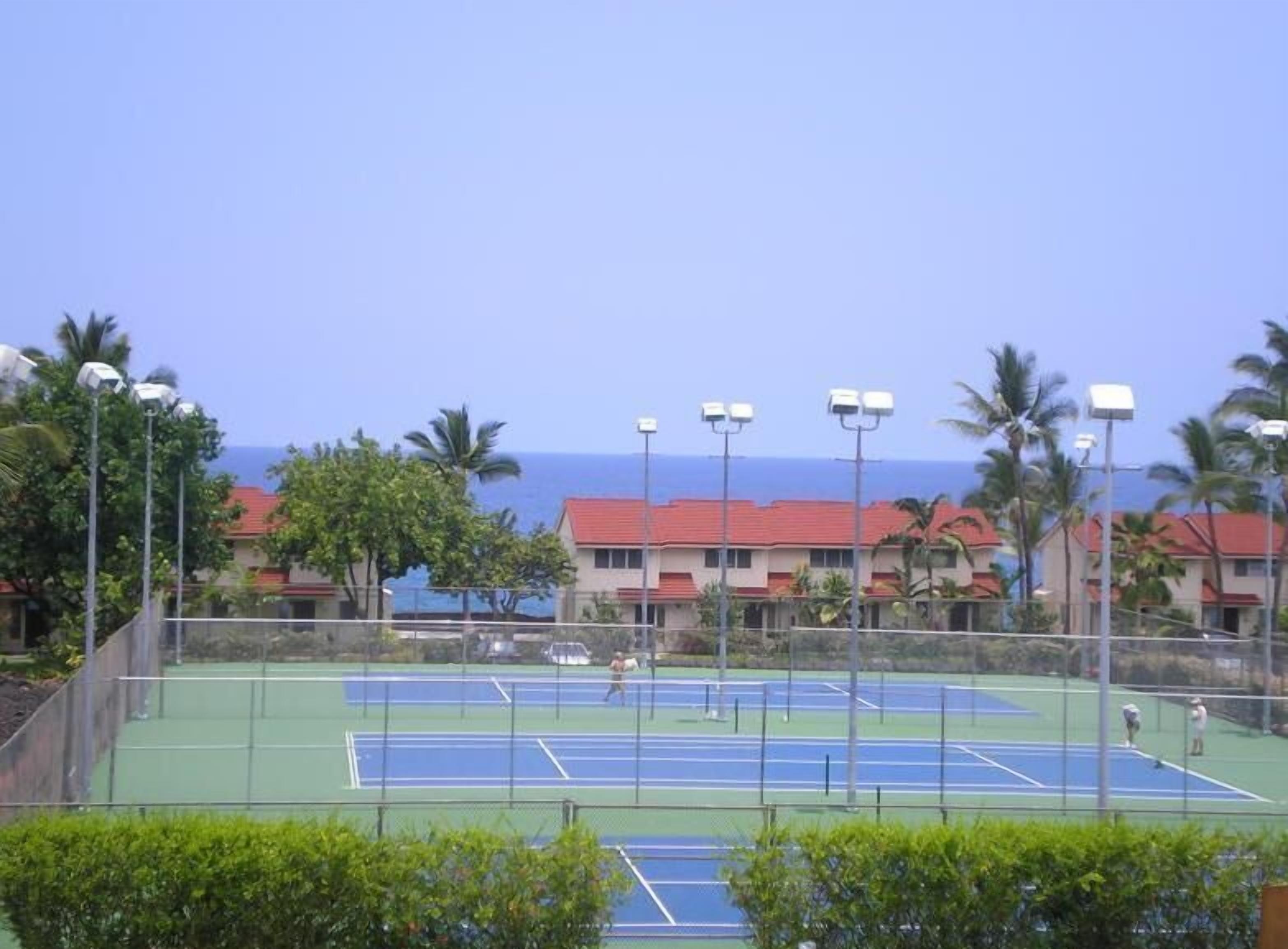 A tennis court with a clear view of the ocean and a row of buildings with red roofs in the background.