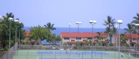 A tennis court with a clear view of the ocean and a row of buildings with red roofs in the background.