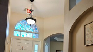 Un couloir avec une porte blanche, un vitrail, un luminaire au plafond et un tableau encadré au mur.