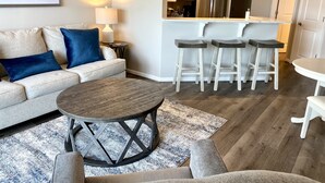 Un salon comprenant un canapé, une table basse et un tapis. Il y a également un coin cuisine avec un réfrigérateur et des tabourets de bar.