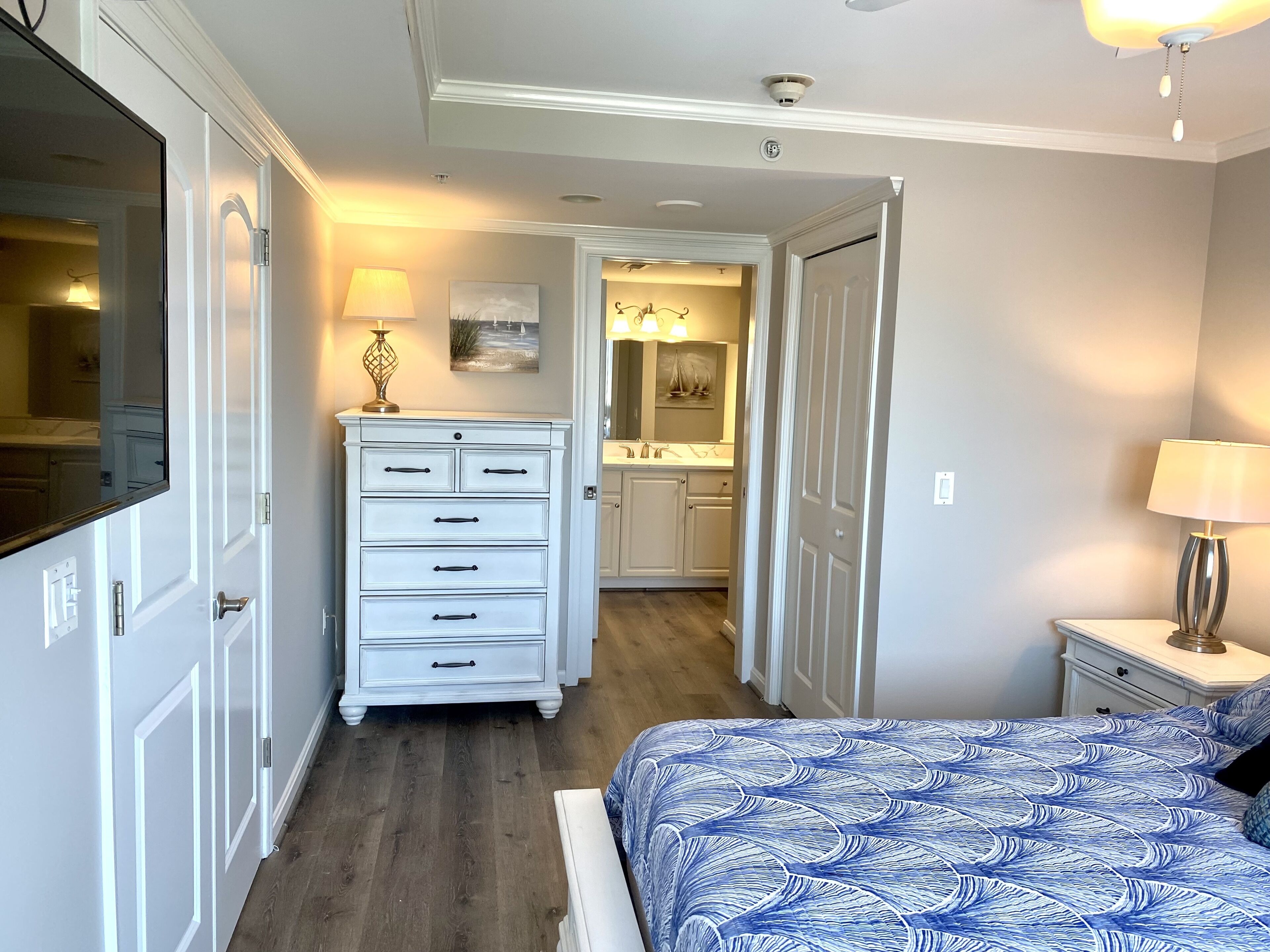 A bedroom with a bed, a dresser, a nightstand, a lamp, and a bathroom visible through an open door.