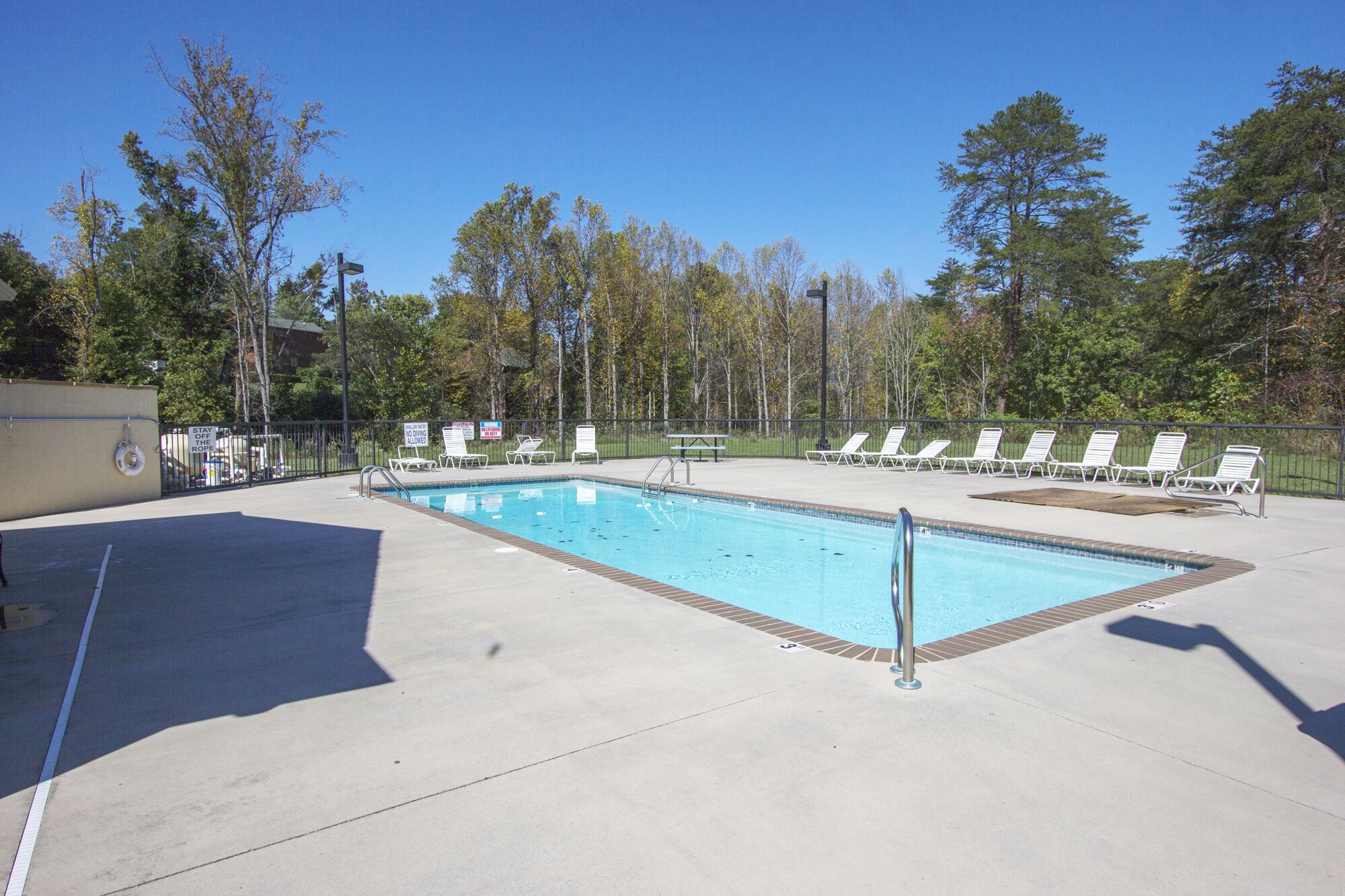 A rectangular pool with a tiled edge, surrounded by a concrete deck. There are white lounge chairs aligned along the pool's edge.