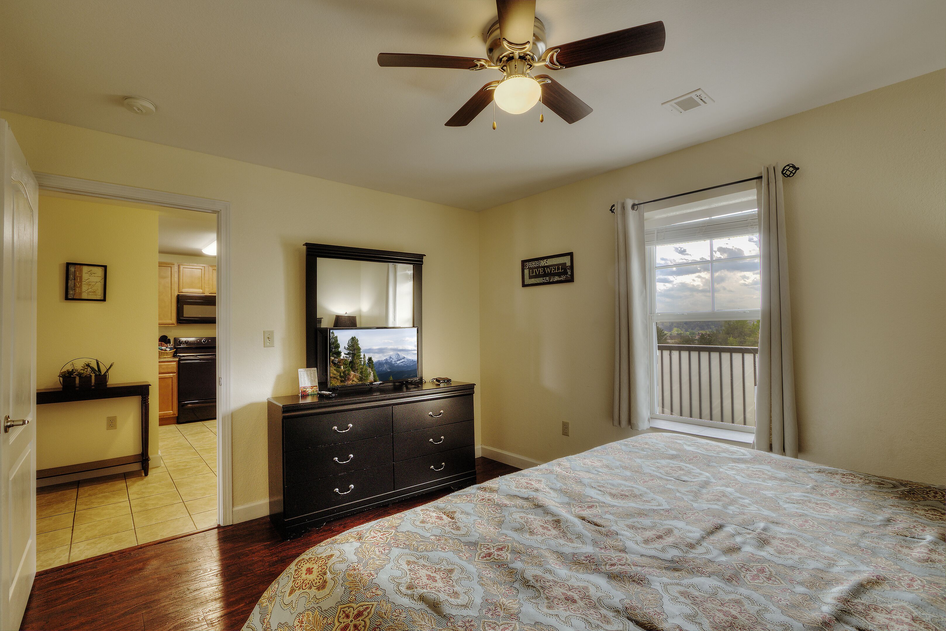 A bedroom with a bed, dresser, and a ceiling fan. There is a window with curtains and a view of the sky.