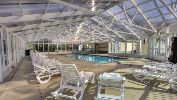 Un área de piscina cubierta con sillas de descanso blancas dispuestas alrededor, una piscina cristalina y una estructura de techo de vidrio.