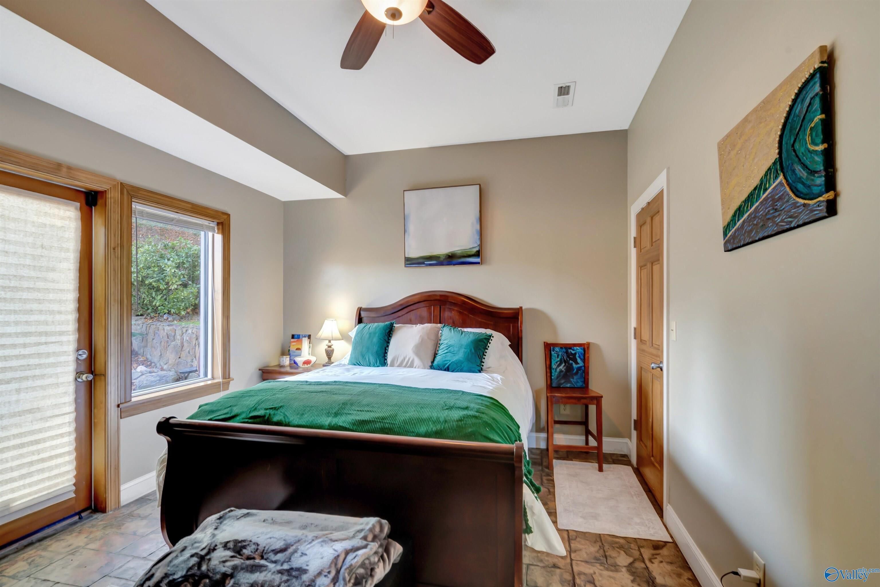 A bedroom with a bed, a ceiling fan, a window with blinds, a painting on the wall, and a door.