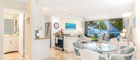 A modern kitchen with white cabinetry, a stainless steel refrigerator, and a built-in oven. There is a dining area with a glass table and white wicker chairs. The room has a large window with a view of palm trees and the ocean.