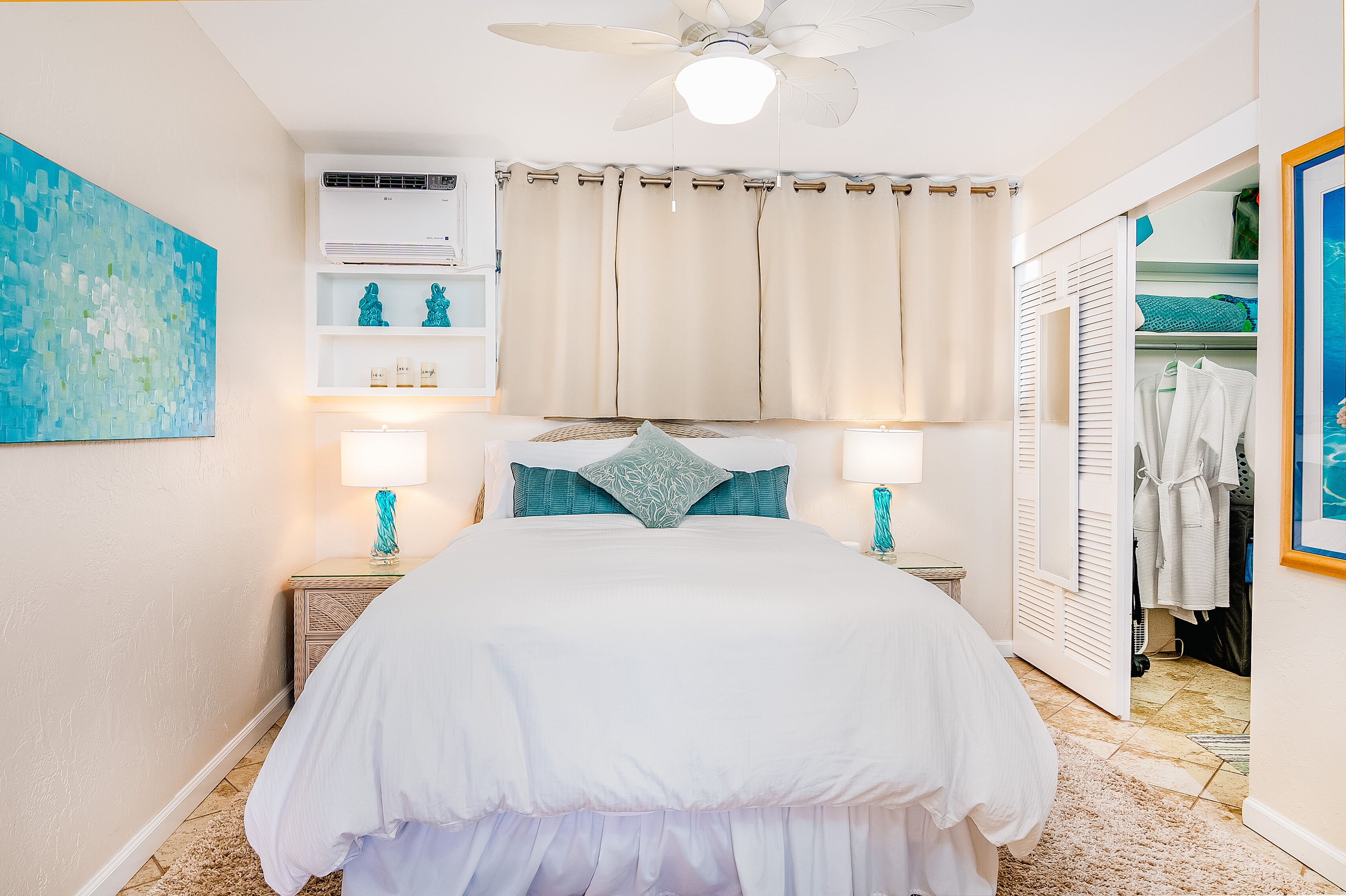 Une chambre à coucher comprenant un lit, deux lampes de chevet, un ventilateur de plafond, un appareil de climatisation, une fenêtre avec des rideaux et une armoire contenant des vêtements suspendus.