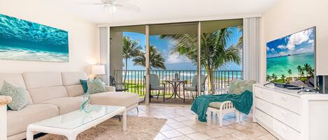 Een moderne woonkamer met een glazen salontafel, een bank en een televisie. De kamer heeft een balkon met uitzicht op zee en palmbomen.