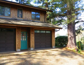 A two-story house with a garage, a green door, and a large tree in the background. - Ocean View Oasis Retreat. Comfort, plus great views! Sleeps 4. (Neskowin)