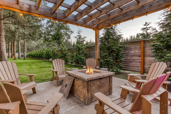 An outdoor patio with a fire pit, wooden chairs, and a pergola with string lights.