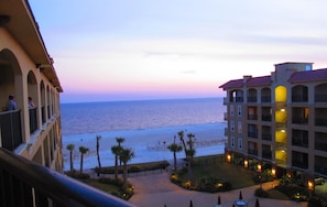 A hotel with a balcony overlooking a beach and ocean. There are palm trees and outdoor seating areas.