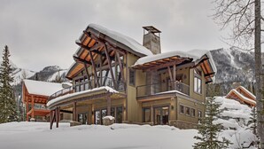 A multi-story lodge with a wooden exterior and a balcony, surrounded by snow-covered trees and mountains.