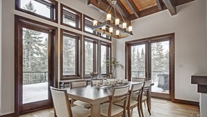 A dining area with a large wooden table and chairs, large windows with a view of snow-covered trees, and a modern chandelier.