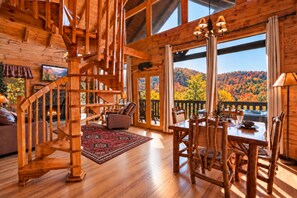 A wooden cabin interior with a dining area, a fireplace, and a view of autumn foliage.