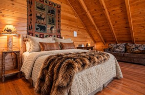A cozy log cabin bedroom with a large bed, a fur throw, a wall tapestry, and a wooden ceiling.