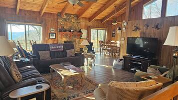 Un intérieur de chalet chaleureux avec une cheminée en pierre, des poutres en bois au plafond, un téléviseur et un coin repas.