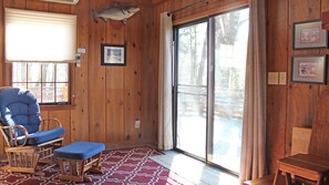 L’intérieur d’un chalet en bois, avec un fauteuil à bascule, un banc et une grande porte-fenêtre coulissante donnant sur une terrasse extérieure.