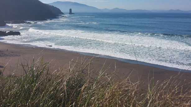 Una playa costera con olas, un acantilado rocoso y montañas a lo lejos.