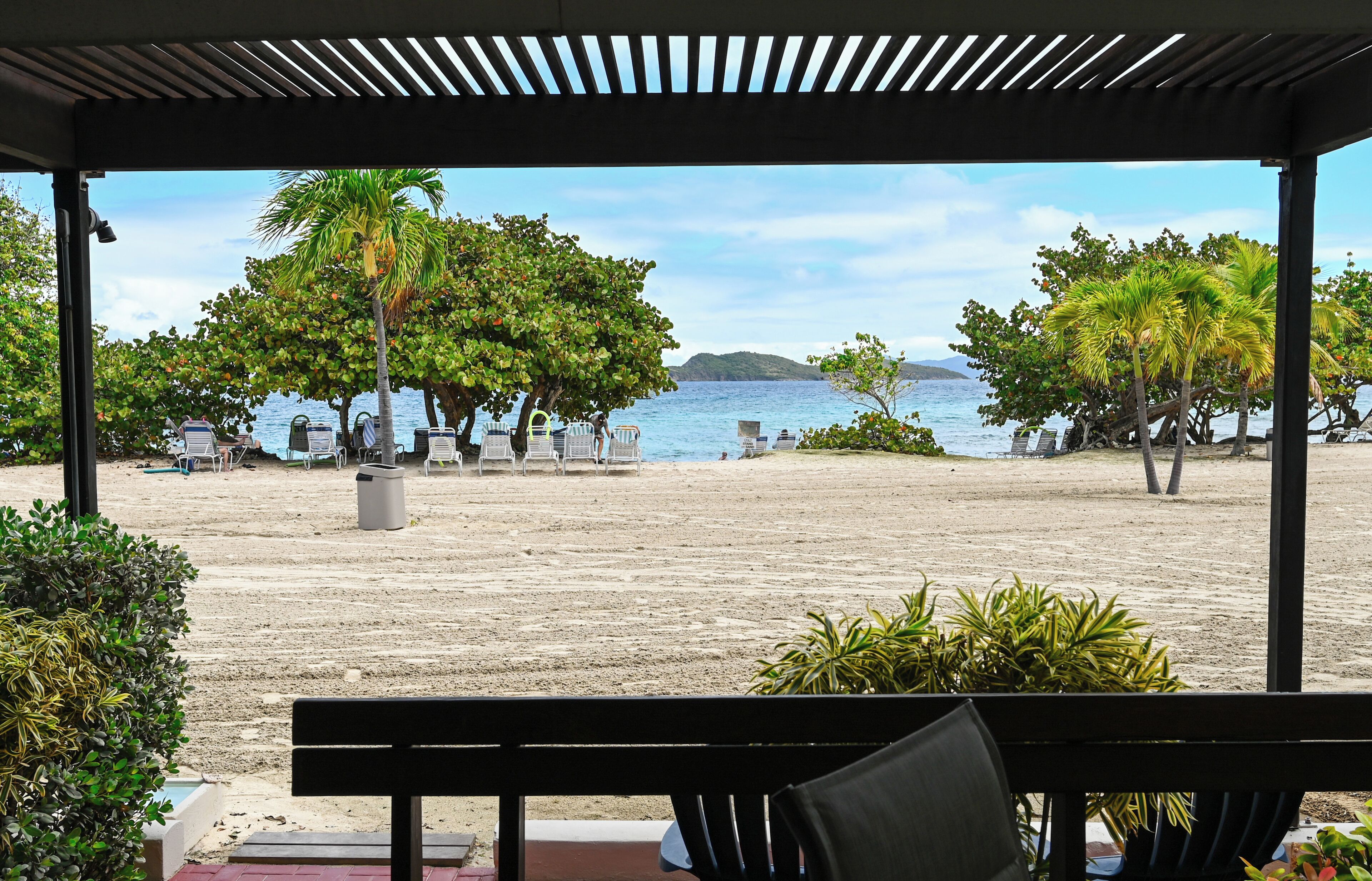 A beach view from a covered area with a bench and chairs, palm trees, and a sandy beach.