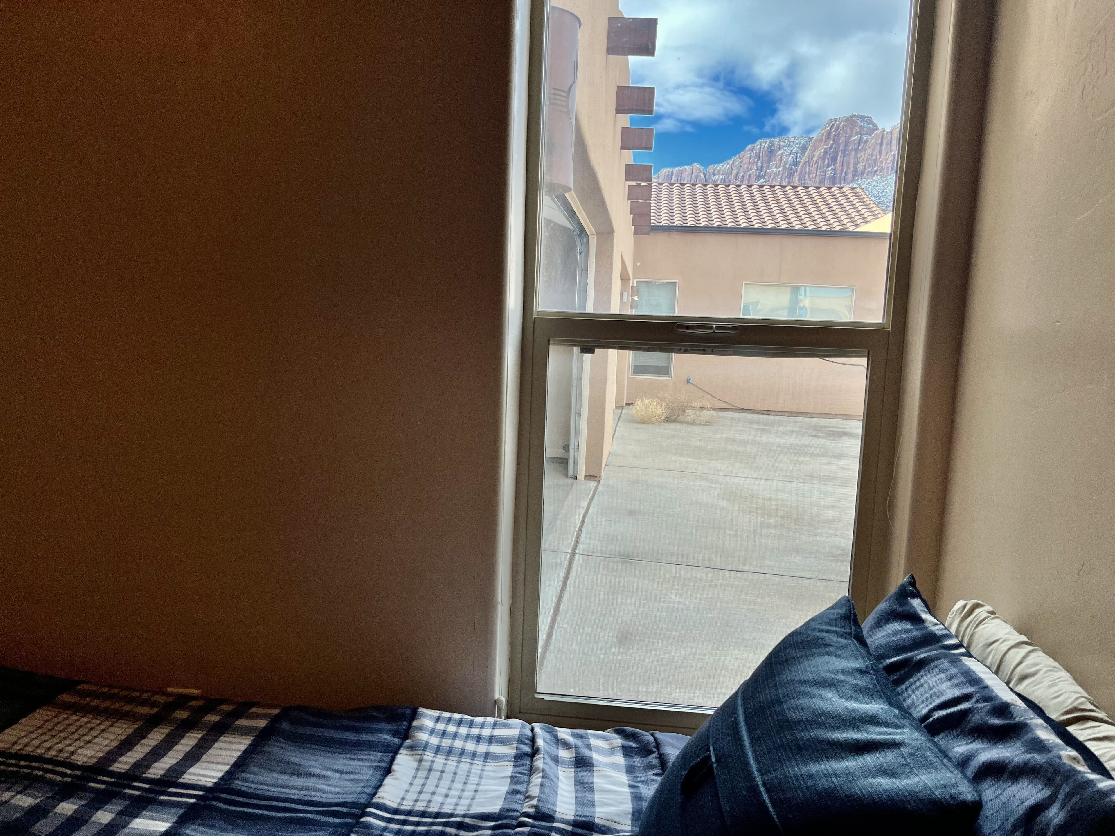 A bedroom with a large window, a bed with a plaid blanket, and a view of a building with a tiled roof.