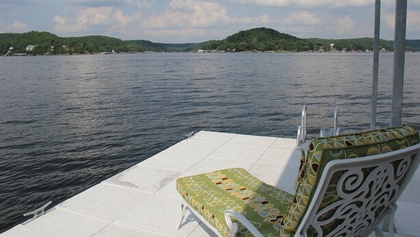 Silla de descanso con cojín estampado en una terraza blanca junto a un lago, con vista a una colina verde y edificios a lo lejos.