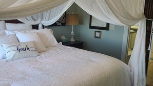 A neatly made bed with white linens and a canopy, a bedside table with a lamp, and a wall with a framed picture.