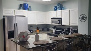 A modern kitchen with white cabinets, a stainless steel refrigerator, a microwave, and a coffee maker. There are four woven placemats on the island, and a sign on the wall that says "Our happy place."
