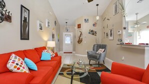 Una sala de estar con un sofá rojo, un sillón gris y una mesa de centro de vidrio. Hay cuadros enmarcados en las paredes y una puerta con un ventanal decorativo.