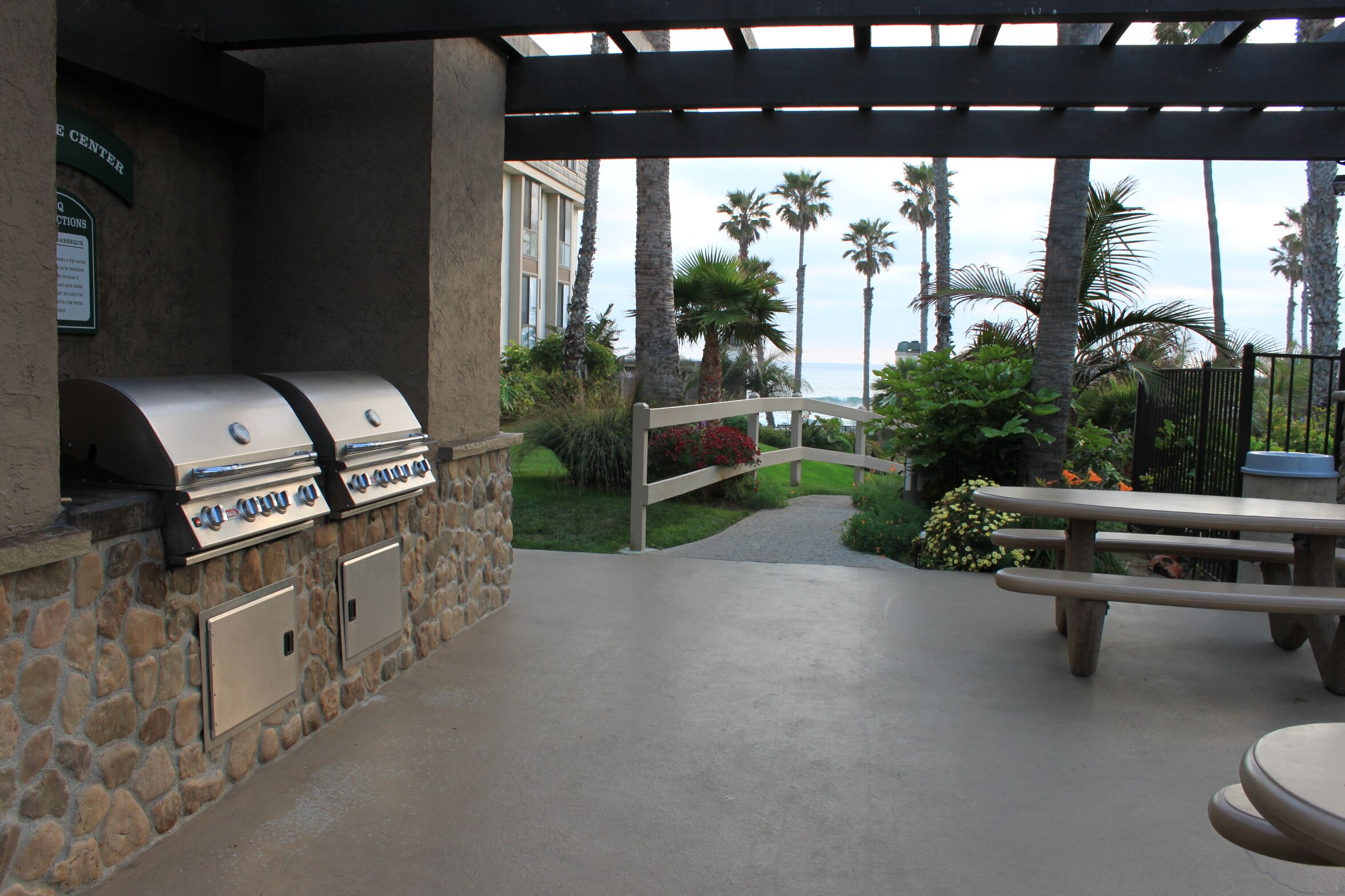 An outdoor patio area with a stone wall, two barbecue grills, a wooden picnic table, and a view of palm trees and a building in the background.