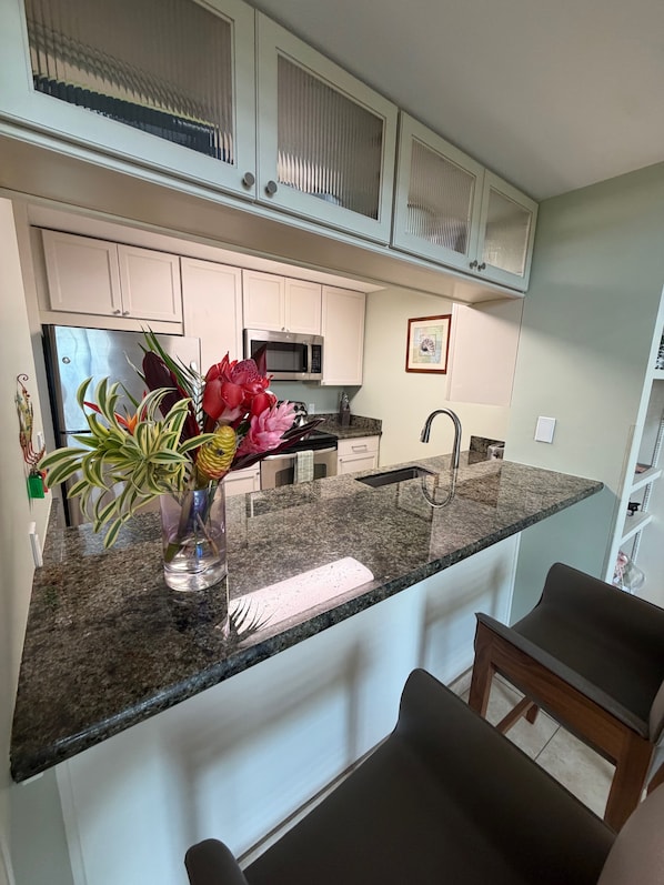 Una cocina con armarios blancos, encimera de granito, refrigerador de acero inoxidable, microondas, pileta y un jarrón con flores.