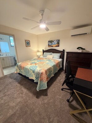 Un dormitorio con una cama, un escritorio, una silla, un ventilador de techo, un cuadro y un equipo de aire acondicionado.