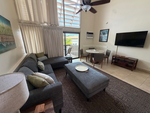 A living room with a sectional sofa, ottoman, dining table with chairs, ceiling fan, TV, and a balcony with curtains.
