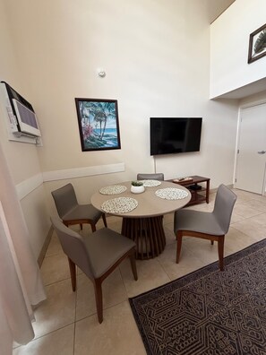 A dining area with a round table, chairs, a TV, and an air conditioning unit.