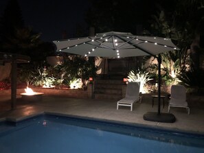A poolside area with a white umbrella, two lounge chairs, and a fire feature.