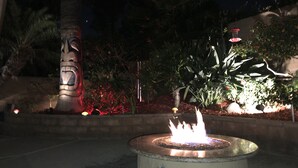 A nighttime outdoor area with a central fire feature, surrounded by various plants and a stone structure.
