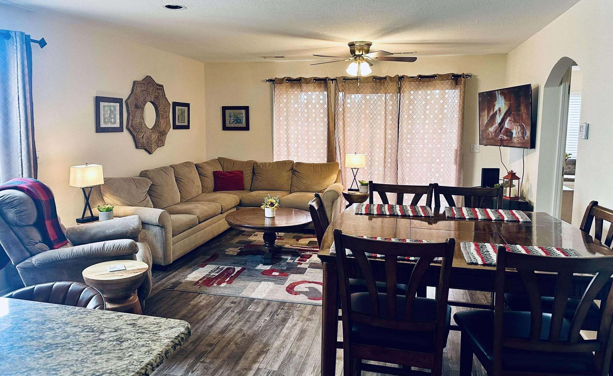 A living room with a sofa, coffee table, dining table, and chairs. There is a ceiling fan, a wall-mounted TV, and a window with curtains.