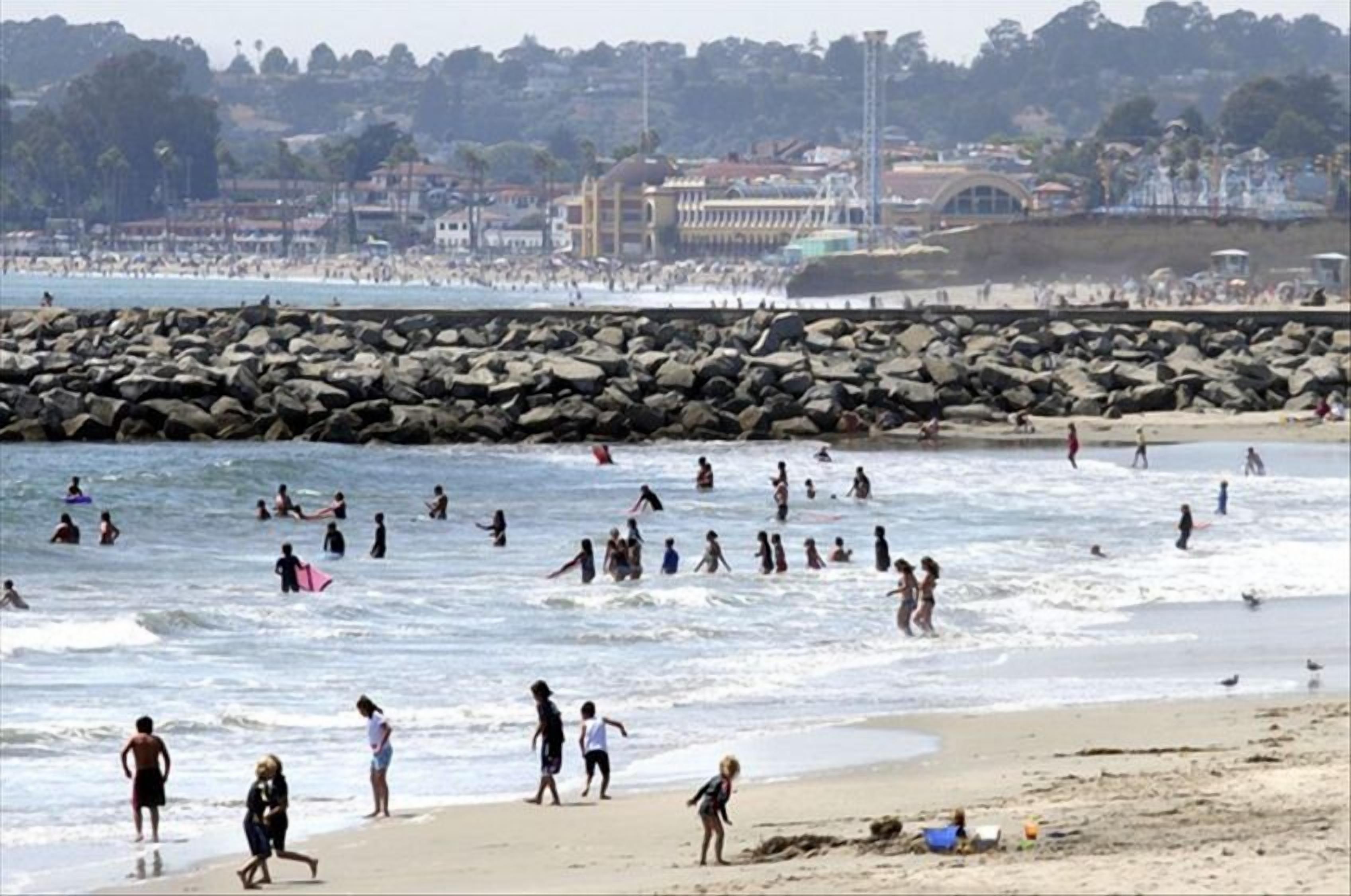 A crowded beach with people swimming and walking along the shore, a rocky breakwater in the background, and buildings on the far side.